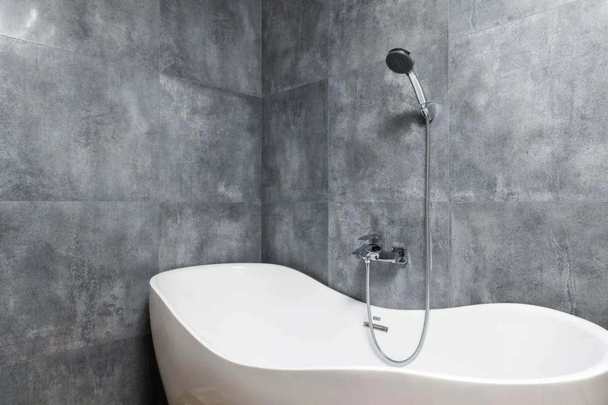 White bathtub under a hand shower against a grey tiled wall