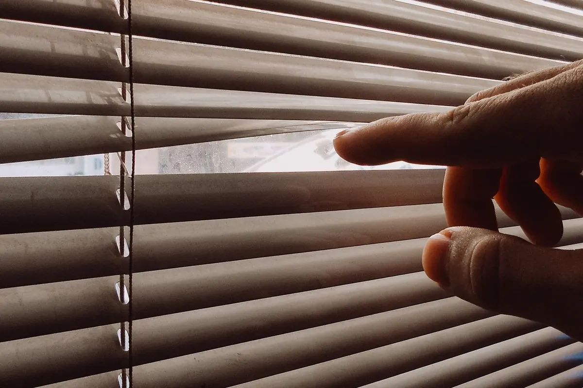 A hand parting the slats of plastic venetian blinds
