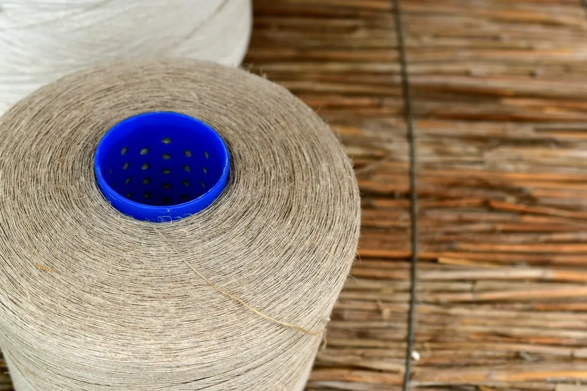 Cone of beige flax yarn on a blue plastic core, resting on a reed mat