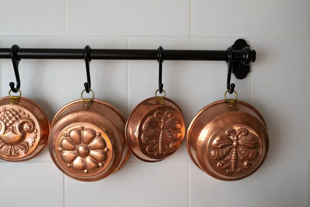 Four red copper baking molds hanging on a rail against a white tiled wall