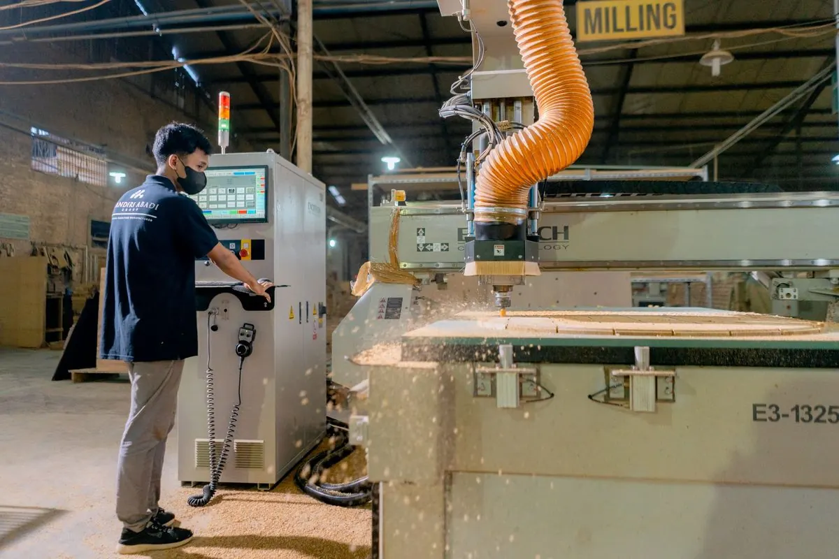 Worker operating a CNC wood router with an orange dust hose