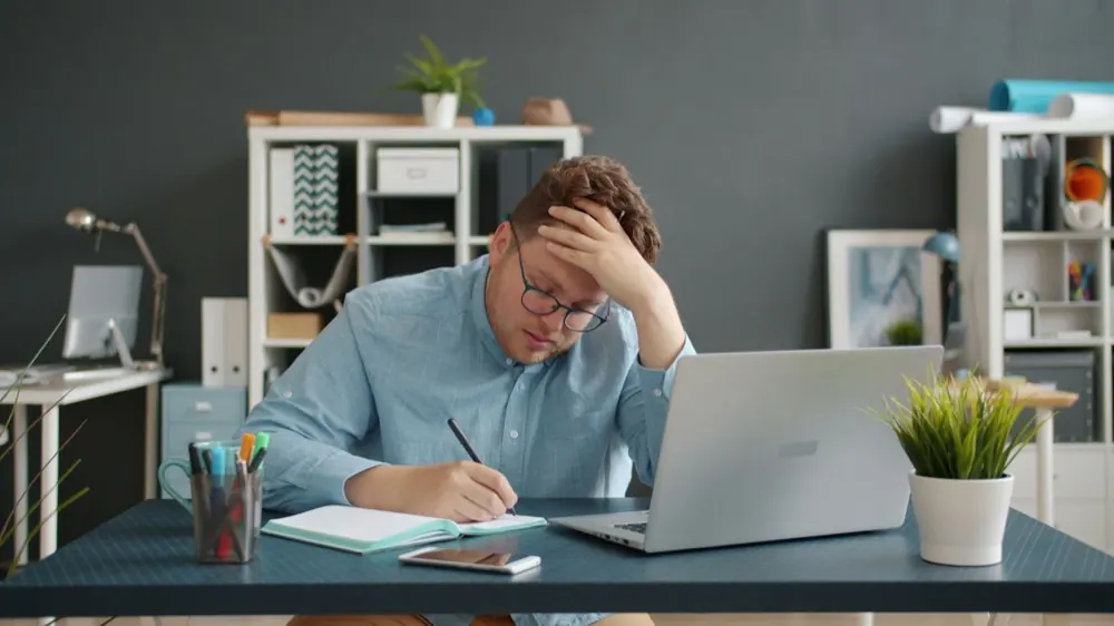 Nhân viên xuất khẩu ngồi trước laptop và sổ ghi chép, tay ôm đầu